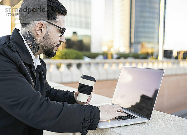 Geschäftsmann  der eine Kaffeetasse hält und einen Laptop an einer Stützmauer benutzt