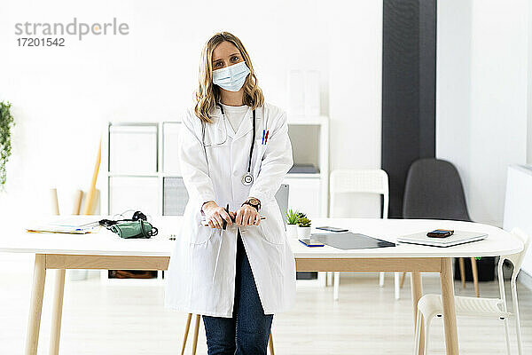 Blonde Ärztin mit Gesichtsschutzmaske und Klemmbrett  die sich im Stehen auf den Schreibtisch stützt
