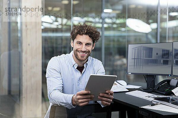 Lächelnder Geschäftsmann  der ein digitales Tablet hält  während er in der Nähe des Schreibtischs im Büro sitzt