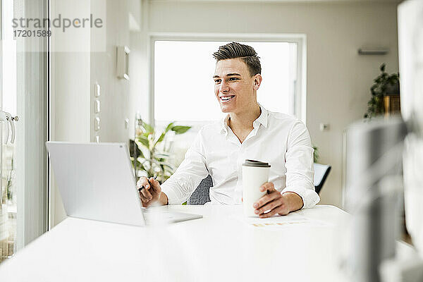 Lächelnder Geschäftsmann mit Einweg-Kaffeebecher und Laptop am Schreibtisch sitzend