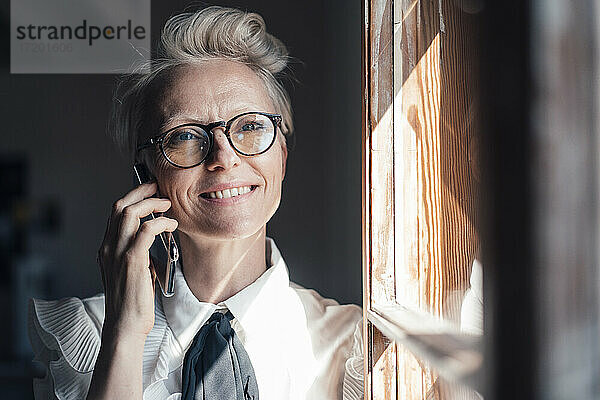Lächelnde Geschäftsfrau mit Brille  die wegschaut  während sie am Fenster im Büro zu Hause mit ihrem Smartphone telefoniert