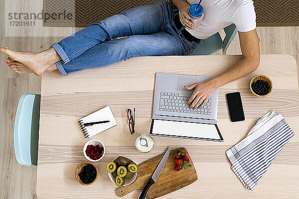 Mann hält Smoothie-Glas  während er am Tisch in der Küche sitzt und einen Laptop benutzt