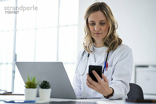 Blonde Ärztin  die ein Mobiltelefon benutzt  sitzt am Schreibtisch in einer medizinischen Klinik