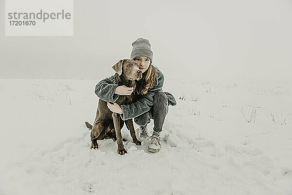 Porträt eines Mädchens im Teenageralter  das im Schnee hockt und einen Labrador Retriever umarmt