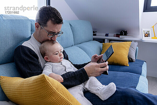 Vater küsst den Kopf seines Sohnes  während er mit seinem Tablet zu Hause auf dem Sofa sitzt