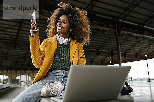 lächelnde Afro-Frau  die ein Mobiltelefon benutzt  während sie vor einem Laptop sitzt