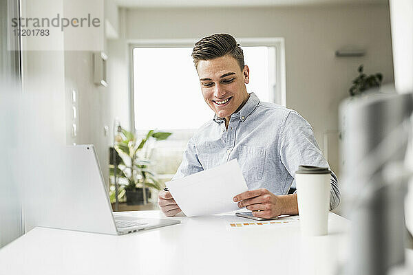 Lächelnder männlicher Fachmann  der ein Dokument liest  während er an einem Tisch im Heimbüro sitzt
