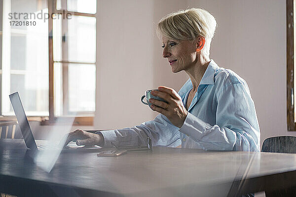 Lächelnde Geschäftsfrau  die eine Kaffeetasse in der Hand hält  während sie an einem Laptop am Schreibtisch im Heimbüro arbeitet