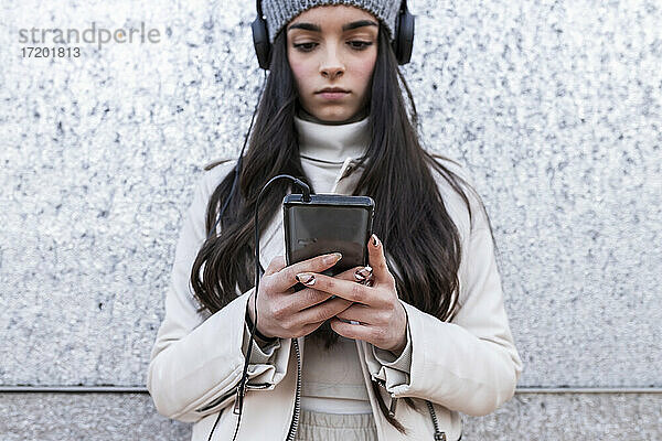 Teenager-Mädchen  das Musik über Kopfhörer hört  während es ein Telefon benutzt  das an einer silbernen Wand steht