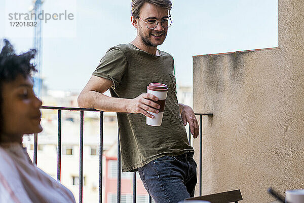 Junger männlicher Unternehmer mit wiederverwendbarem Becher  der während einer Kaffeepause im Büro am Geländer lehnt