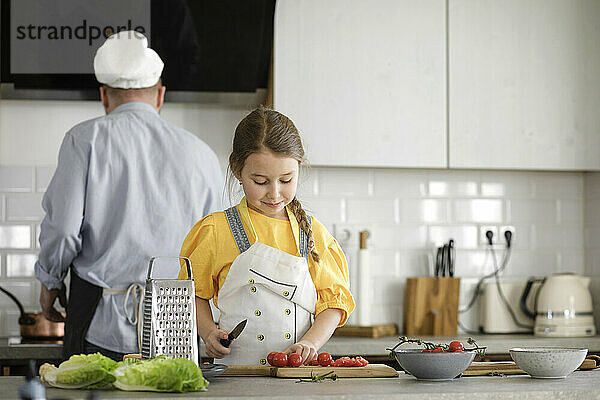 Mädchen  das Tomaten schneidet  während es in der Küche steht  mit Vater im Hintergrund zu Hause
