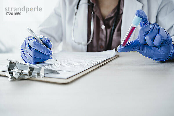 Eine Ärztin hält eine Blutprobe und schreibt einen medizinischen Bericht am Schreibtisch