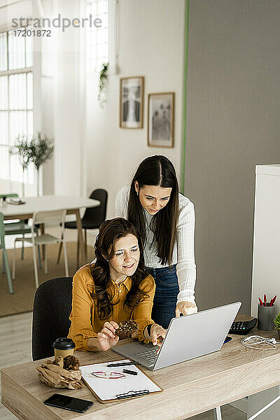 Geschäftsfrau mit Tochter  die auf einen Laptop schaut  während sie einen Keks am Schreibtisch im Heimbüro isst