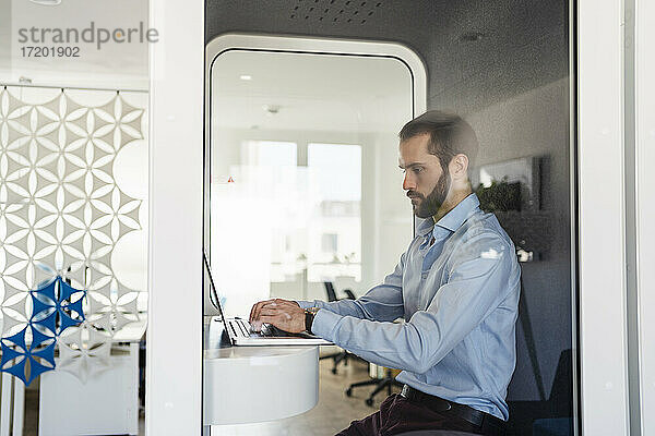Männlicher Berufstätiger arbeitet am Laptop  während er in einer Telefonzelle im Büro sitzt