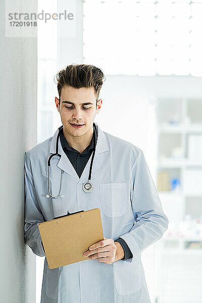 Männlicher Arzt schreibt Notizen  während er sich im Krankenhaus an die Wand lehnt