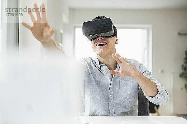 Fröhlicher männlicher Berufstätiger  der einen Virtual-Reality-Simulator trägt  während er am Schreibtisch im Heimbüro sitzt