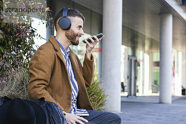 Geschäftsmann mit Kopfhörern  der mit seinem Mobiltelefon spricht  während er mit einer Tasche im Freien sitzt