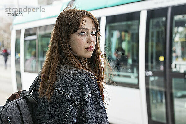 Junge Frau mit braunen Haaren steht an einer Straßenbahnhaltestelle vor einem Zug