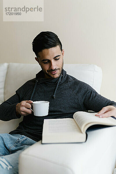 Junger Mann mit Kaffeetasse  der zu Hause auf dem Sofa sitzt und ein Buch liest