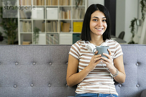 Glückliche Frau mit Kaffeetasse und kabellosen Kopfhörern auf dem Sofa zu Hause