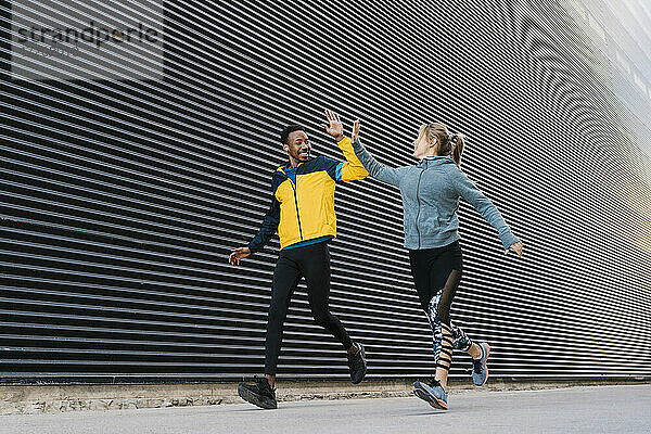 Männlicher und weiblicher Sportler machen High-Five beim Laufen auf dem Gehweg während des Sporttrainings an der Wand