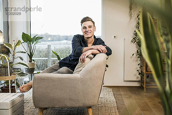 Lächelnder junger Mann sitzt auf dem Sofa vor dem Fenster im Wohnzimmer