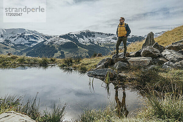 Mann mit Händen in den Taschen steht auf einem Felsen vor einem Teich im Salzburger Land. Leoganger Berge  Österreich