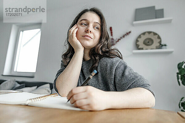 Nachdenkliche Frau sitzt am Tisch und schreibt in ein Notizbuch im Wohnzimmer