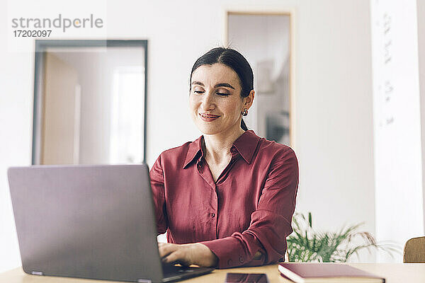 Lächelnde Unternehmerin  die einen Laptop benutzt  während sie am Tisch im Heimbüro sitzt