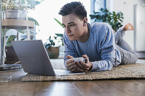 Junger Mann hält Kaffeetasse  während er ein Video auf dem Laptop ansieht