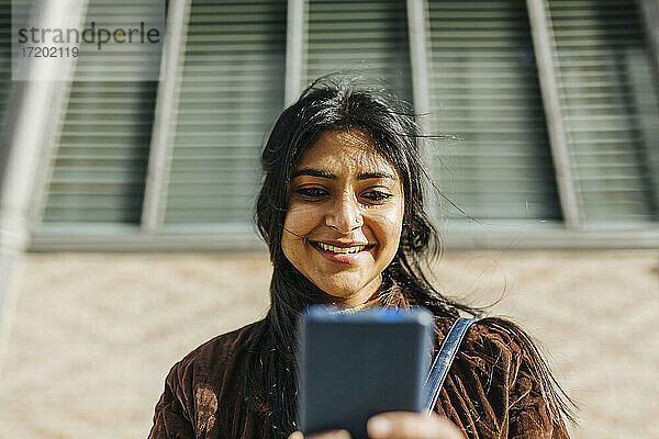 Lächelnde Frau  die ein Mobiltelefon gegen ein Gebäude benutzt