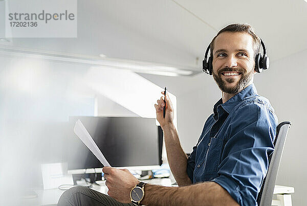 Lächelnder Geschäftsmann mit Kopfhörern und Papier im Büro sitzend