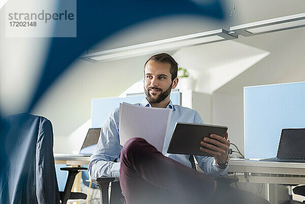 Unternehmer mit digitalem Tablet und Papier  der lächelnd im Büro sitzt