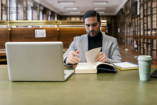 Mittlerer Erwachsener Geschäftsmann mit Laptop  der in einer Bibliothek sitzt und ein Buch liest