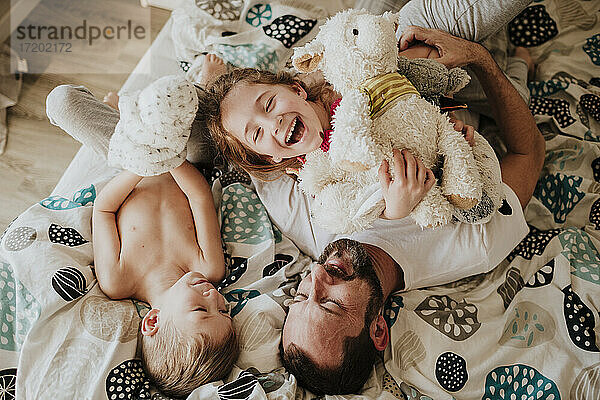 Lachender Vater und spielende Tochter  während sie mit ihrem Sohn zu Hause auf dem Bett liegen