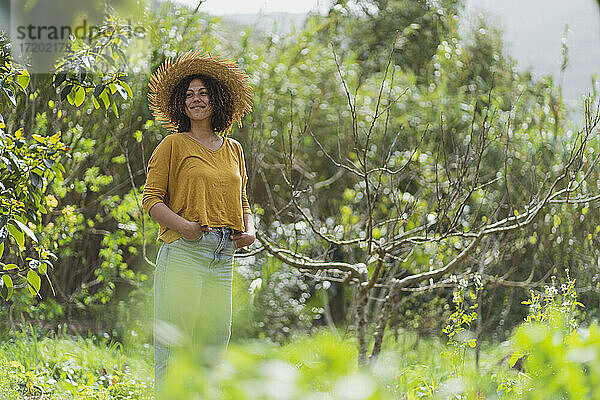 Lächelnde Frau mit Strohhut und Händen in den Taschen steht an einem kahlen Baum im Garten