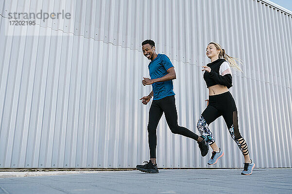 Lächelnder männlicher und weiblicher Athlet  der während des Sporttrainings auf dem Gehweg an einer Mauer läuft