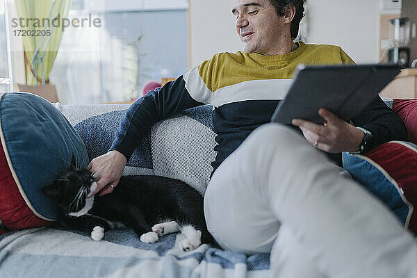 Lächelnder Mann mit digitalem Tablet  der seine Katze streichelt  während er auf dem Sofa im Wohnzimmer sitzt