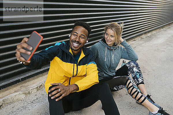 Fröhlicher Sportler mit Sportlerin  die ein Selfie mit ihrem Handy macht  während sie an der Wand sitzen