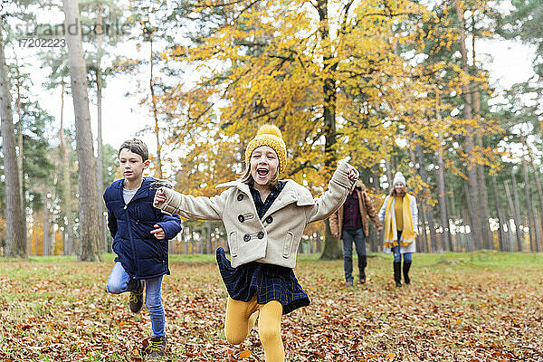 Mädchen und Junge laufen beim Spielen mit Eltern im Hintergrund im Wald