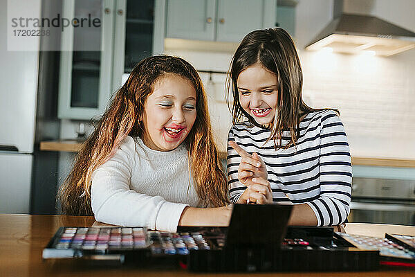 Fröhliche Schwestern spielen mit Make-up-Palette  während sie am Esstisch in der Küche sitzen