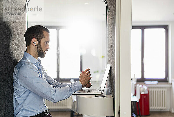 Geschäftsmann  der an einer Besprechung am Laptop teilnimmt  während er in einer Telefonzelle im Büro sitzt