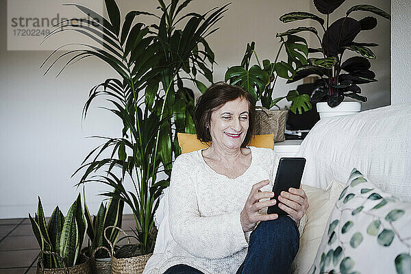 Lächelnde Frau  die ein Mobiltelefon benutzt  während sie zu Hause auf dem Sofa sitzt