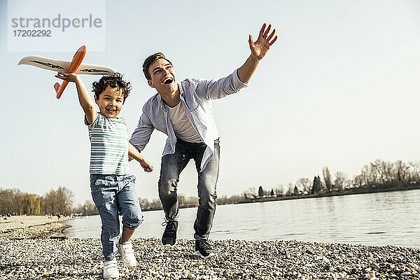 Verspielter Mann und Junge halten Flugzeugspielzeug  während sie am Seeufer an einem sonnigen Tag laufen