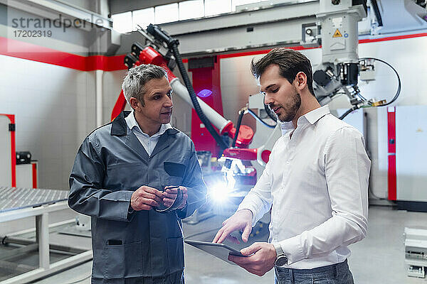 Junger männlicher Ingenieur  der ein digitales Tablet benutzt  während er sich mit einem reifen Mann in einer Fabrik unterhält