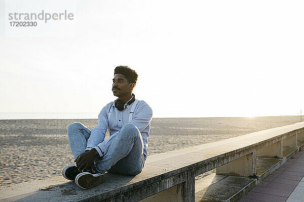 Nachdenklicher Mann sitzt auf einer Stützmauer am Strand gegen den klaren Himmel an einem sonnigen Tag
