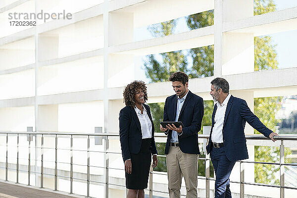 Ein professionelles Team diskutiert über ein digitales Tablet  während es an einem Geländer steht.