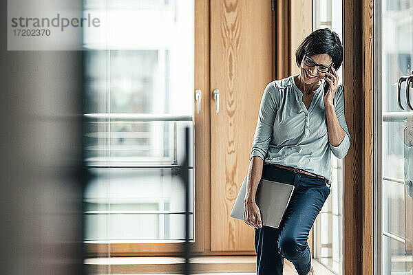 Unternehmerin  die mit ihrem Smartphone telefoniert  während sie sich im Büro über das Fenster lehnt