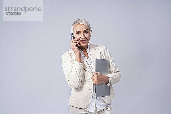 Lächelnde Geschäftsfrau  die mit einem Mobiltelefon spricht  während sie vor einem grauen Hintergrund steht