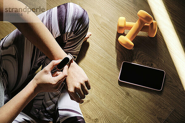 Junge Frau  die eine intelligente Uhr benutzt  während sie zu Hause im Schneidersitz auf dem Boden sitzt